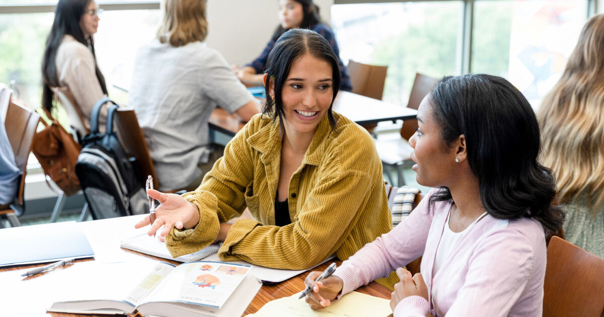 Two teacher candidates sit next to each other in one of their educator preparation classes. They are talking to one another and taking notes.