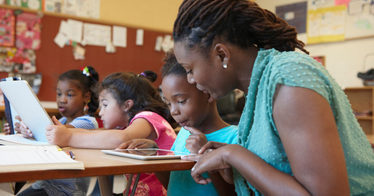 African American female teacher in a classroom helping a young student at her desk.