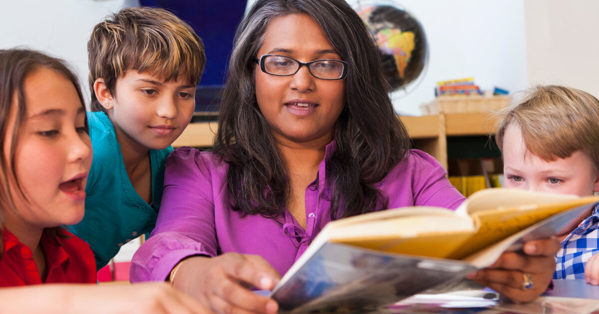 In a classroom, a female teacher reading a book to 3 students